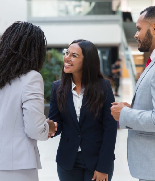 Female diverse business partners shaking hands with each other in hallway. Business man and women standing in office hall, talking and smiling. Successful partnership concept